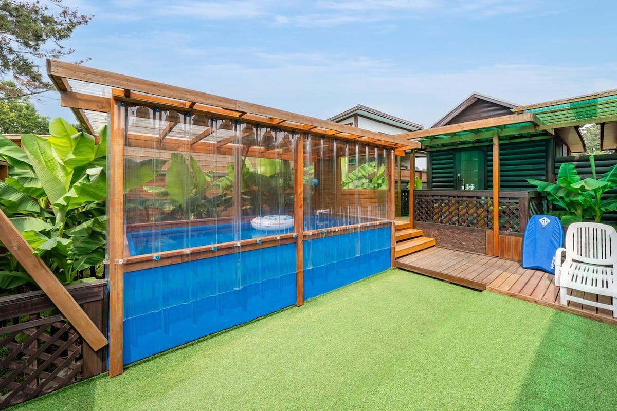 A backyard with a swimming pool, artificial grass, wooden deck, and a covered seating area.