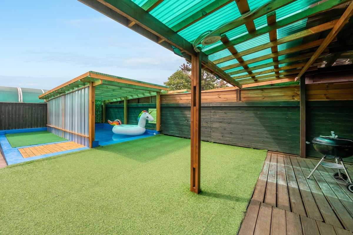 A backyard with artificial turf, a wooden deck, a covered play area, and a barbecue grill.