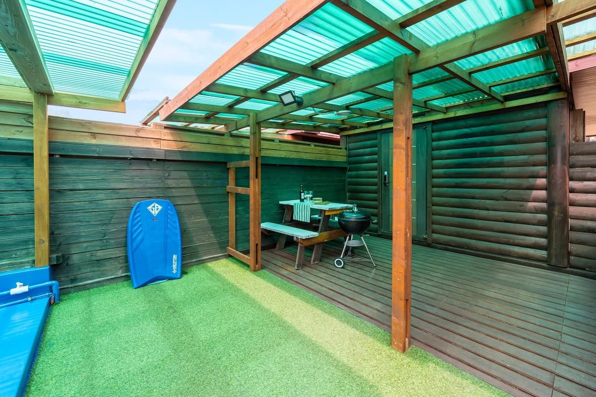 A covered outdoor area with a wooden deck, a barbecue setup, and a blue sauna.