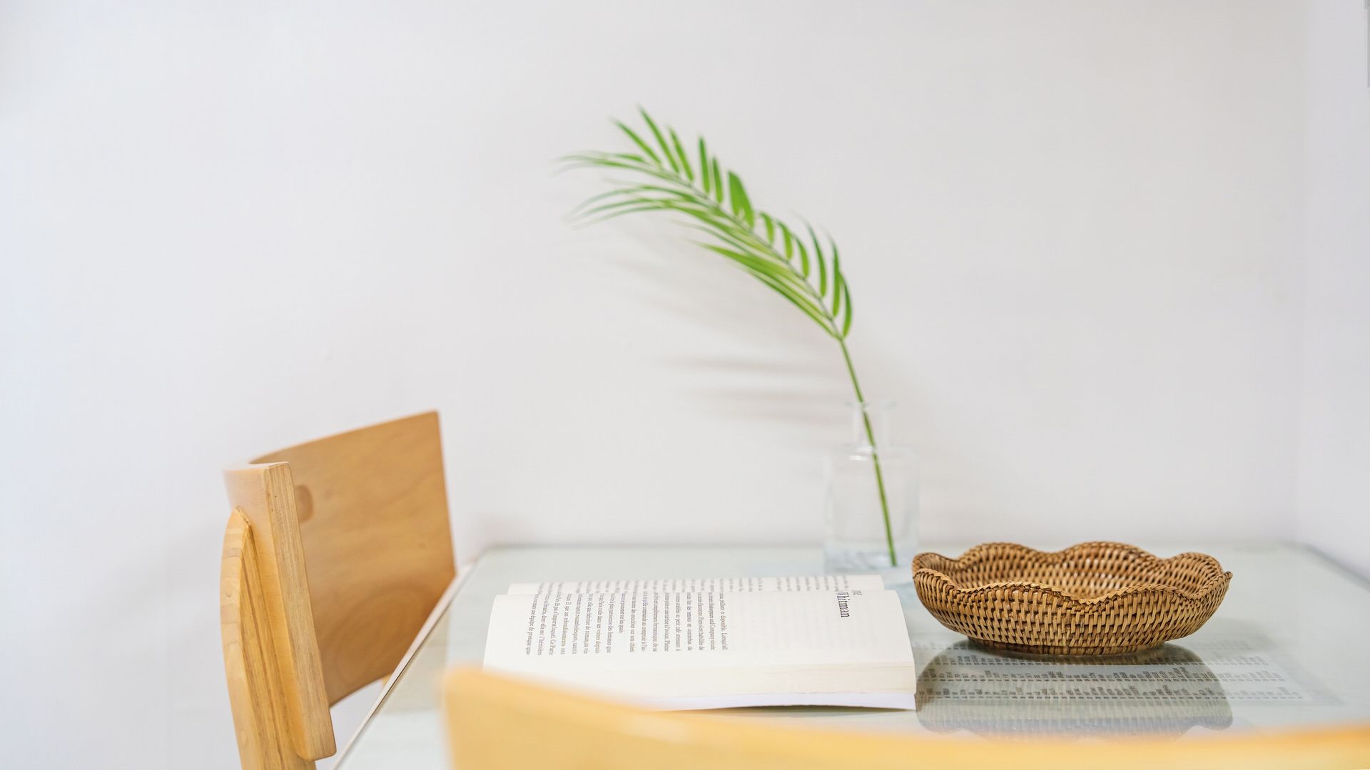 Un serre-livre en bois, un livre et un panier tressé sont posés sur une table, à côté d’un vase en verre transparent contenant une seule plante verte.