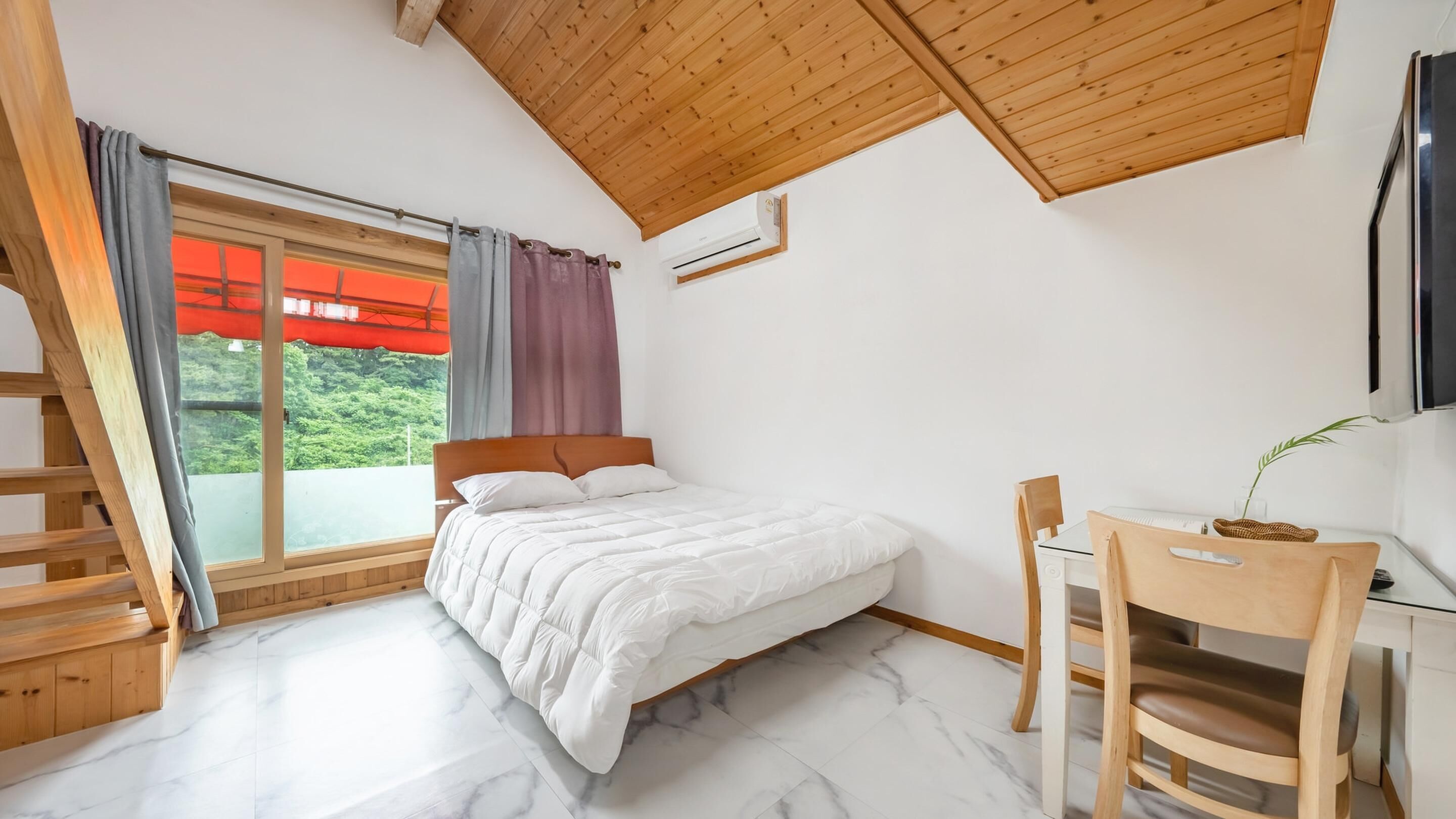 A bedroom with a bed, a desk, and a chair. There is a window with curtains and a view of greenery outside.