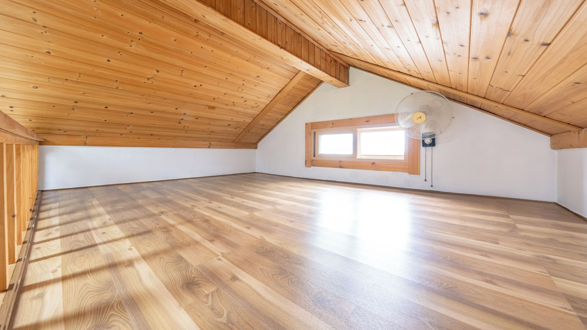 Une chambre mansardée avec un plancher en bois, un plafond en pente et une fenêtre surmontée d’un tympan.
