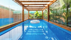 Una piscina interior con cerramiento de cristal transparente, vigas de madera y un flotador.