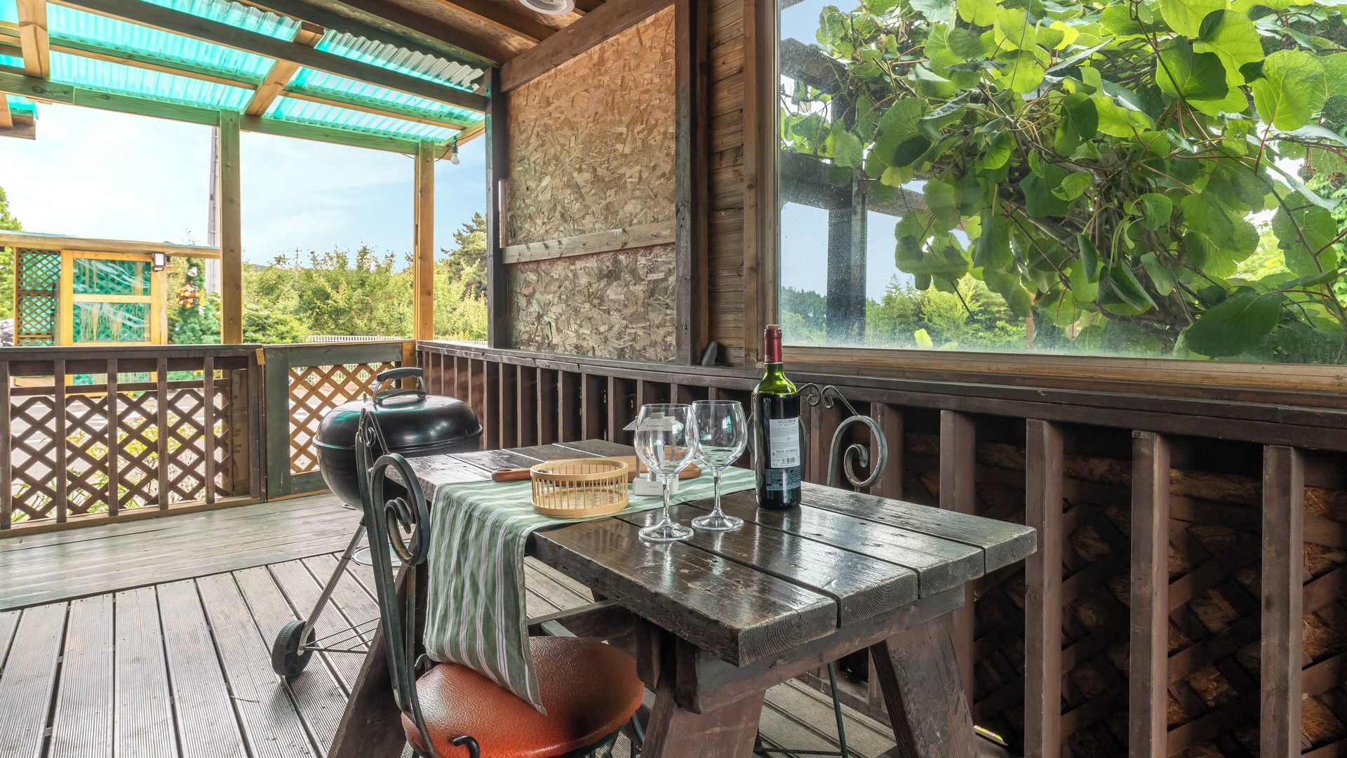An outdoor wooden deck with a table set for dining, a grill, and a view of greenery.