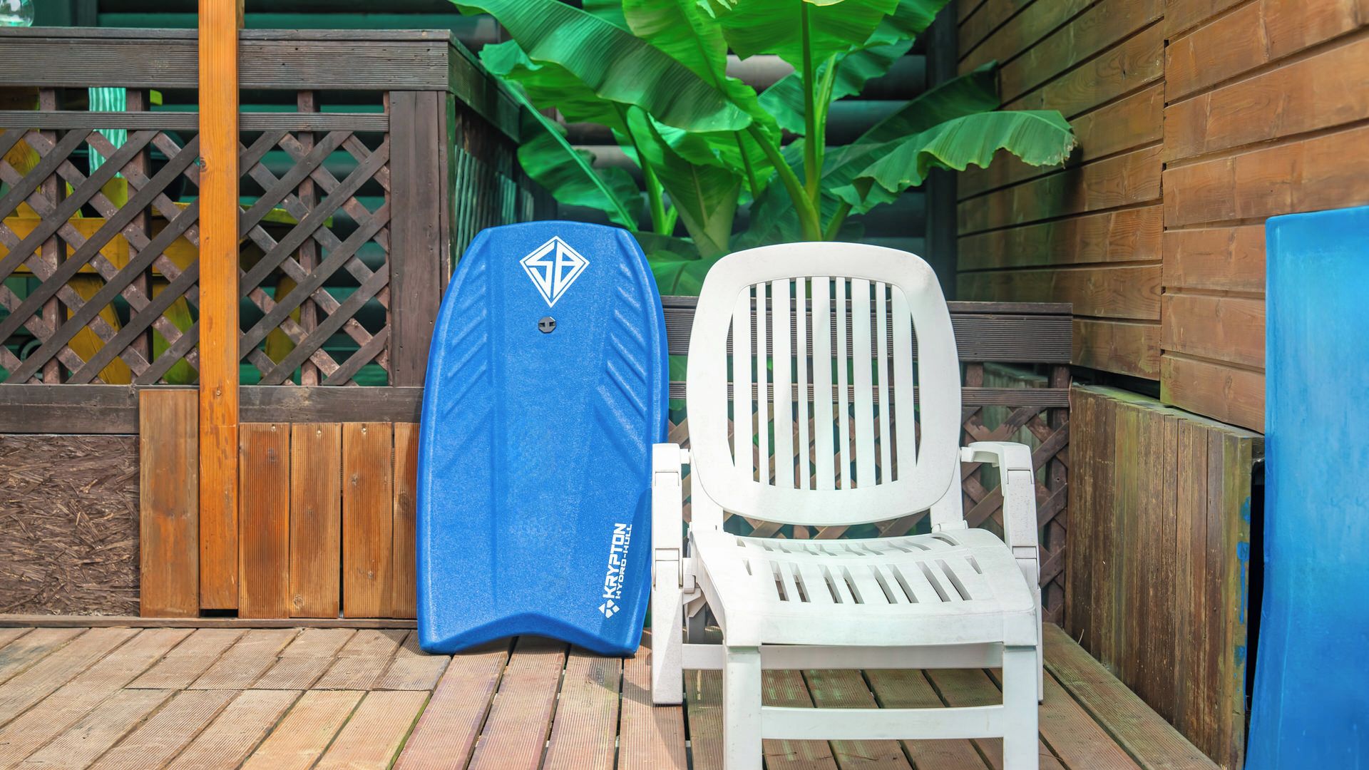 A white lounge chair with a blue cover, a wooden fence, and a large green plant in the background.