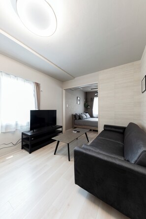 A modern living room with a flat-screen TV on a black stand, a black sofa, and a coffee table. There is a wooden floor and a large window with curtains. - NEMOPHILA ALPHA S7W8 106 (Sapporo)