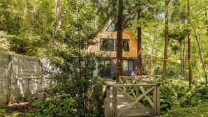 Una casa de madera rodeada de un bosque tupido, con un puente de madera que lleva a la entrada.