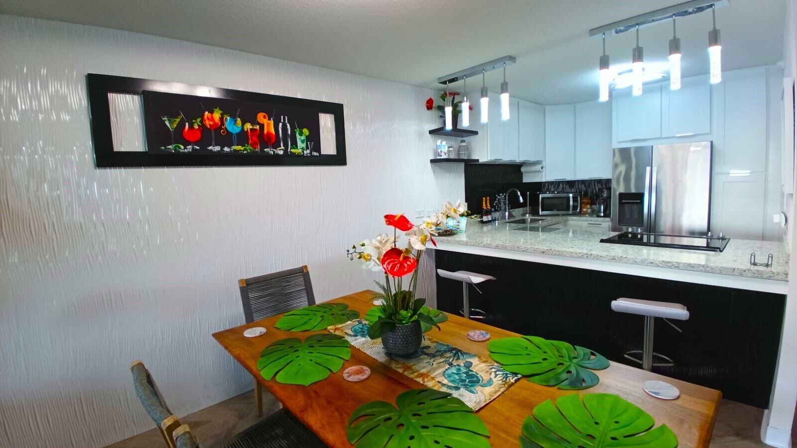 A modern kitchen with a central island, a dining table with a tropical leaf-patterned table runner, and a colorful artwork above the island.