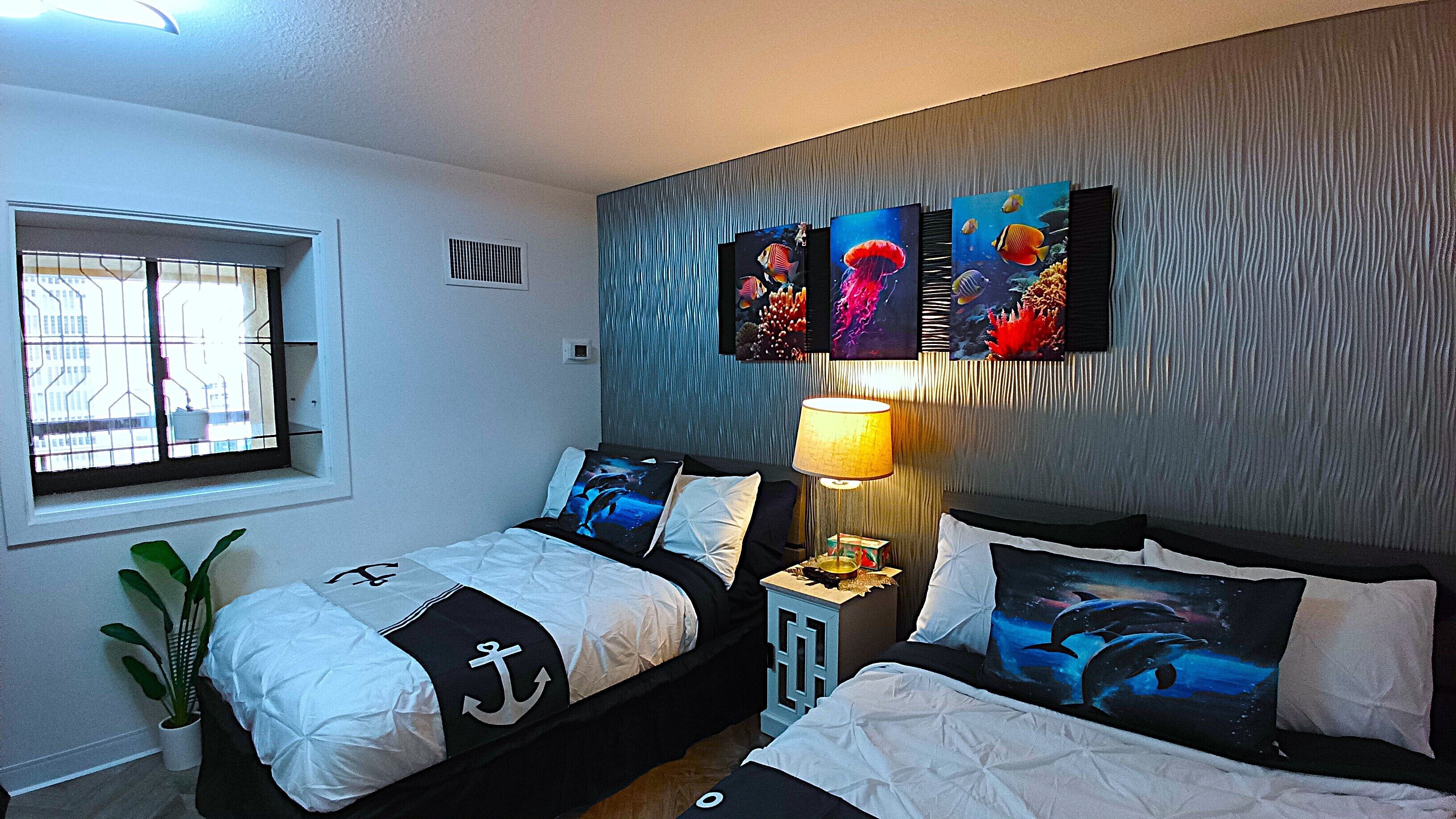 A bedroom with two beds, a window, a plant, and three paintings on the wall.