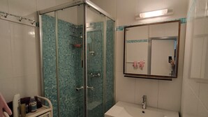 Un bany amb dutxa trencada per vidre, un lavabo blanc i un mirall a sobre.