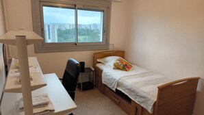Habitació petita i ordenada amb llit, escriptori i cadira. Hi ha una finestra amb vista a la ciutat, i l'espai té un disseny senzill i net.
