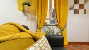 A room with a sofa, yellow curtains, a window with blinds, a wall-mounted plant, and a framed informational poster. - Happy Family Apartments Andreana (Genoa)