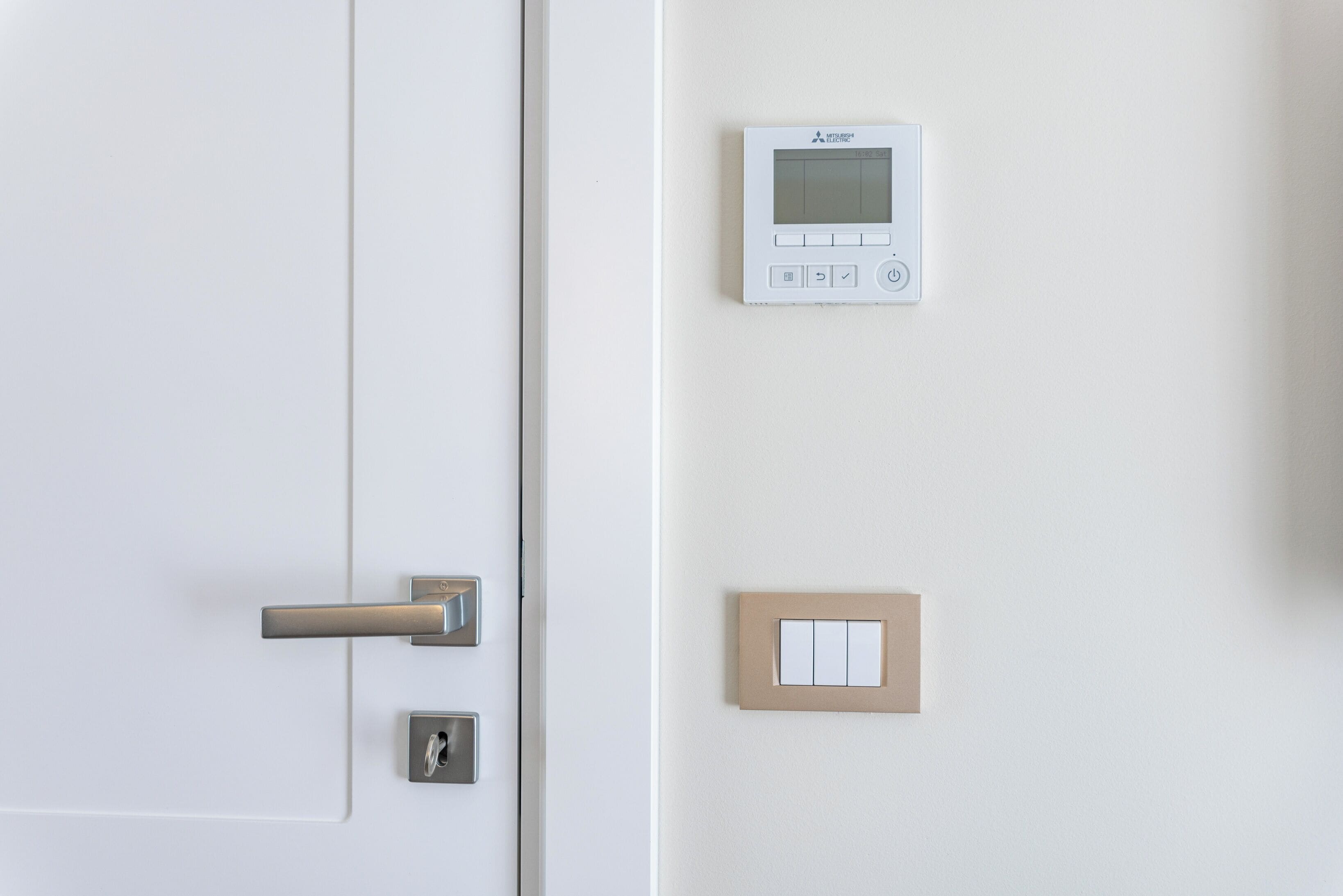 Una puerta blanca con manija plateada y un ojo de cerradura, un termostato instalado en la pared y un interruptor de luz.