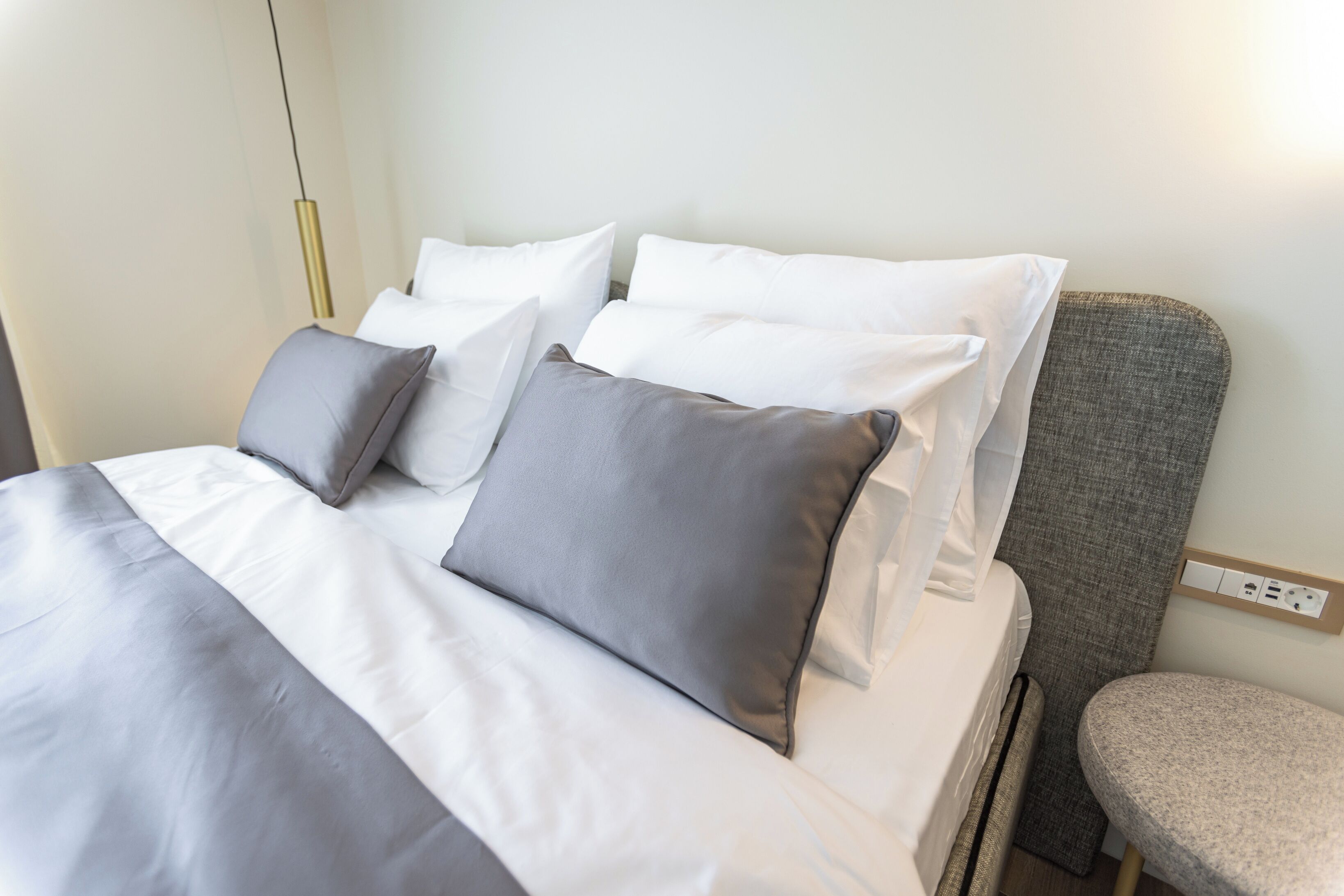Una cama bien hecha con ropa de cama blanca y gris, un cabecero tapizado en gris y una pequeña mesa redonda junto a la cama.