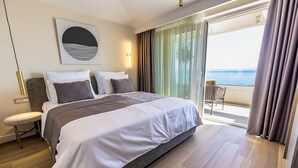 A modern hotel room with a large bed, a seating area with a chair, and a balcony with a view of the sea.