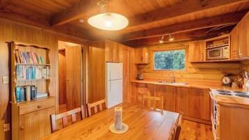 A wooden cabin interior with a dining table, bookshelves, a refrigerator, a microwave, and a window overlooking greenery.