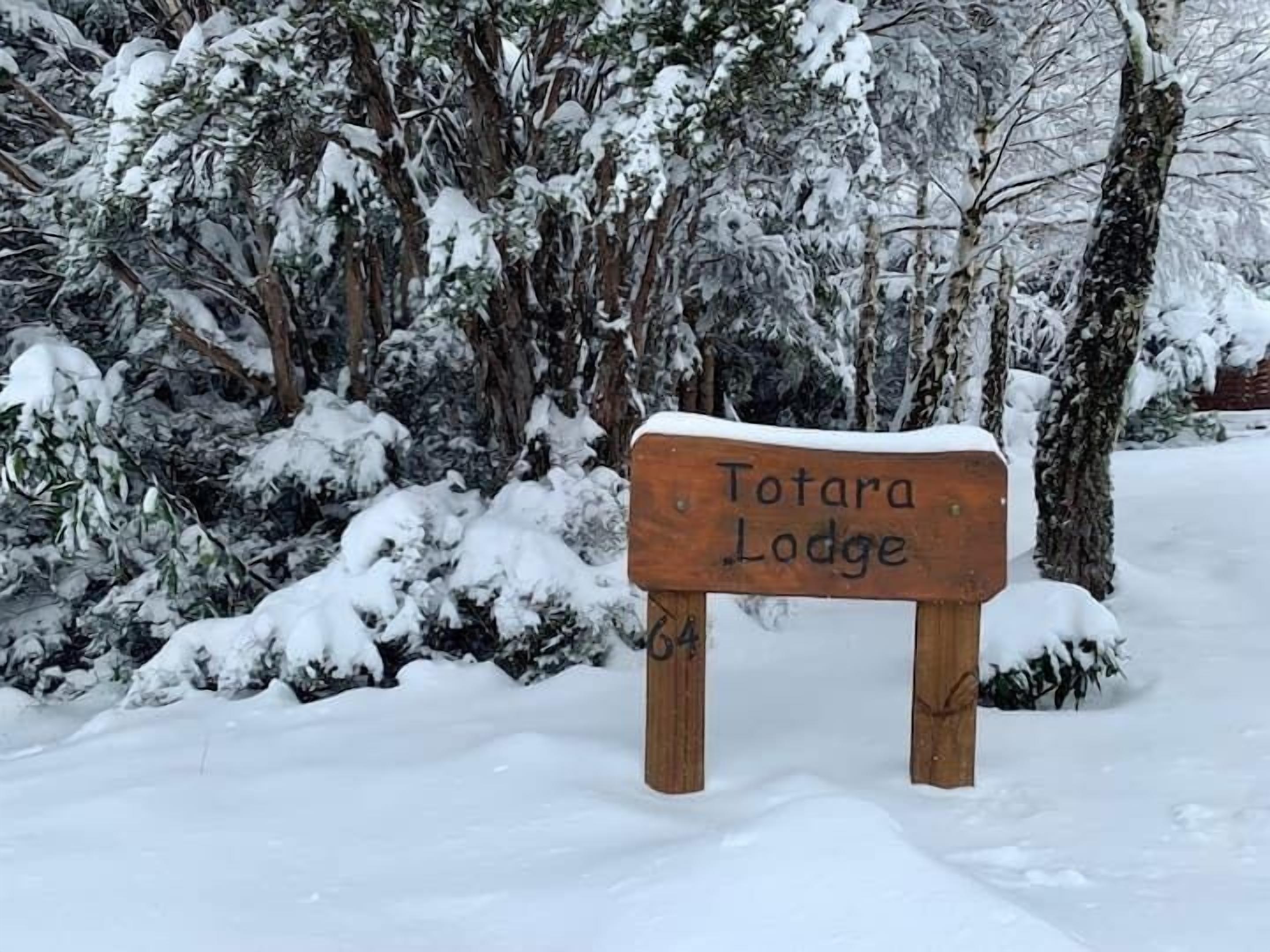 A wooden sign with 'Totara Lodge' written on it, surrounded by snow-covered trees.