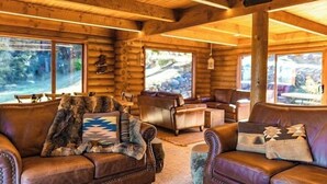 Interior de una cabaña de troncos con sofás de cuero, una chimenea y amplias ventanas que ofrecen vistas del exterior.