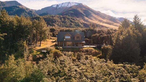 Una casa con techo inclinado, rodeada de un bosque denso y montañas al fondo.