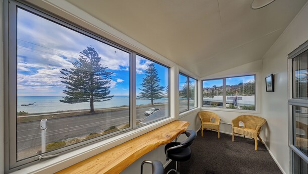 A room with large windows offering a view of the sea and trees, a wooden bench, two wicker chairs, and a small table. - Waterfront | Handy Location | Affordable | Unit 1 - KK41164 (Kaikoura)