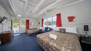 A hotel room with a large bed, a smaller sofa, a desk, and a TV. There is a window with red curtains and a painting on the wall. - Waterfront | Handy Location | Affordable | Unit 1 - KK41164 (Kaikoura)