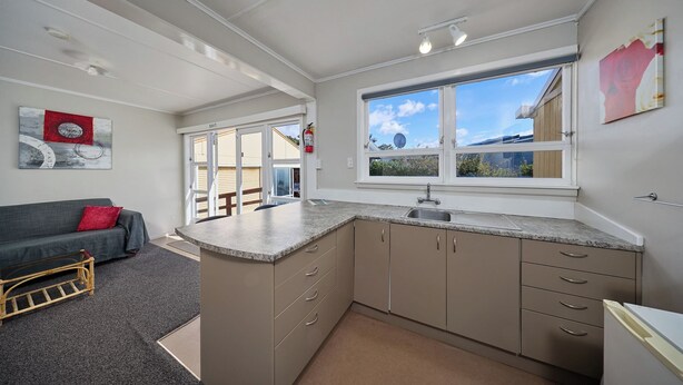 A compact kitchen with a built-in island, a dining area with a grey sofa and a small wooden table, and a large window offering a view of the outside. - Waterfront | Handy Location | Affordable | Unit 6 - KK61646 (Kaikoura)