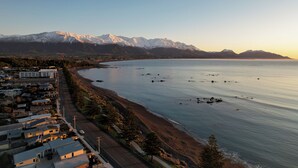 A coastal town with a beach, a road lined with buildings, and snow-capped mountains in the background. - Waterfront | Handy Location | Affordable | Unit 6 - KK61646 (Kaikoura)