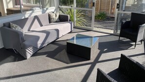 Uma área de estar moderna ao ar livre com um sofá cinzento, uma mesa de centro de vidro e cadeiras de vime.