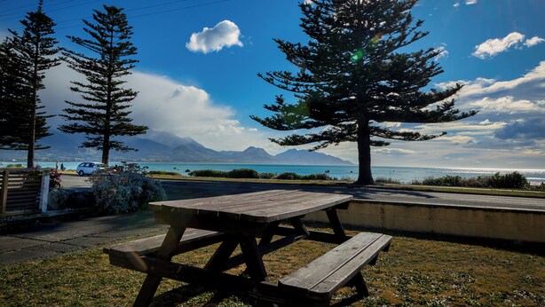 Uma mesa de piquenique com bancos, vista para um lago e montanhas ao fundo.