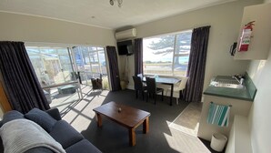 Uma sala de estar moderna com sofá, mesa de centro e área de jantar com cadeiras e mesa. Há uma janela com cortinas, um extintor de incêndio e uma pia pequena.