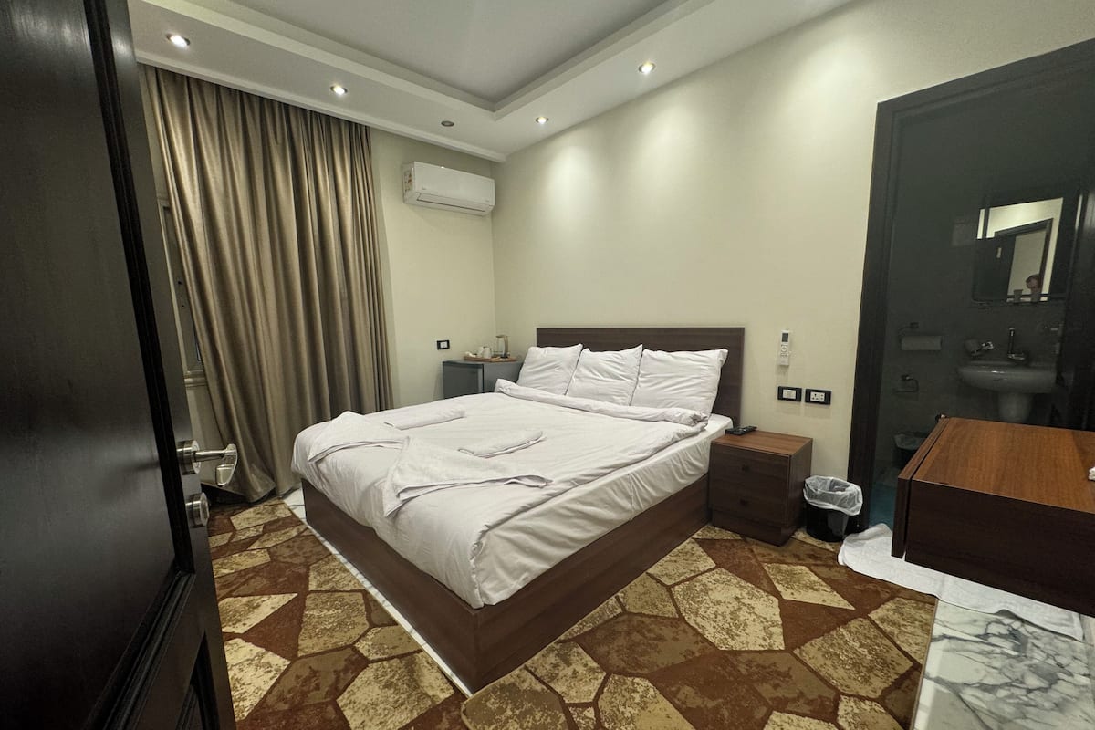 A hotel room with a large bed, a bedside table, a nightstand, a bathroom with a sink and mirror, and a patterned floor.