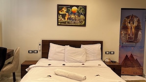 Un lit bien fait, avec du linge de lit blanc, une tête de lit et un oreiller. Il y a deux tables de chevet avec des lampes et une œuvre d’art accrochée au mur représentant une scène avec des palmiers et un soleil.