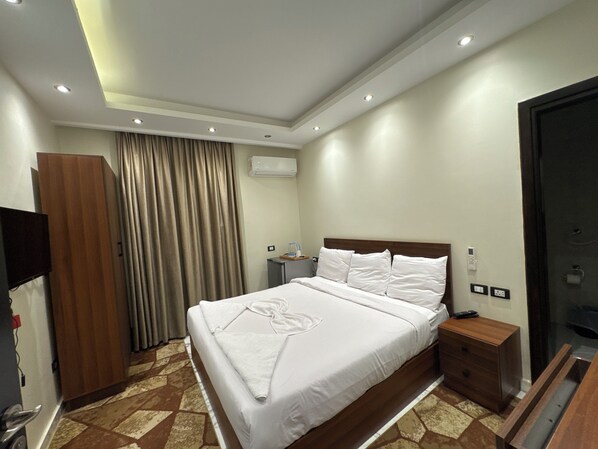 A hotel room with a large bed, a wooden wardrobe, an air conditioning unit, a bedside table with a lamp, and a television on the wall. - Unique pyramids view inn (Giza)
