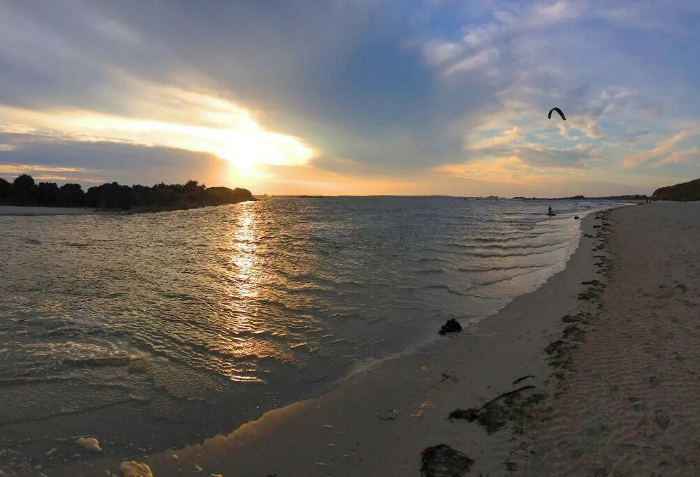 Ein Strand mit Sonnenuntergang, ruhigem Wasser und einer Person, die ein Drachen steigen lässt.