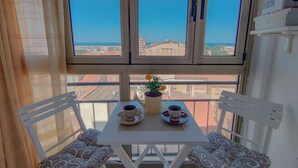 Ein Balkon mit Tisch, zwei Stühlen, einem Topfpflanzen und Blick auf Gebäude und Meer.