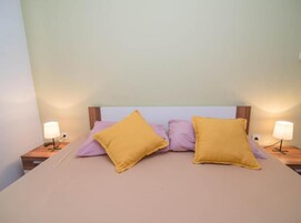 Una cama con dos almohadas amarillas y dos moradas, flanqueada por dos mesitas de noche con lámparas.