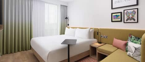 Camera d'albergo con un letto, comodino, lampada e un divano. Si vedono tende, una parete con quadri e una finestra.