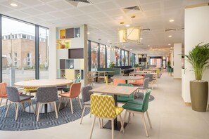 A modern interior with round tables and colorful chairs, large windows, and a view of the outside. - Hampton By Hilton Nuneaton (Nuneaton)