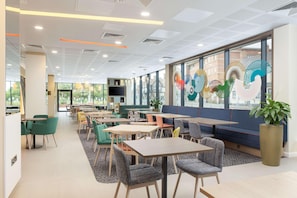 A modern dining area with tables and chairs, large windows, and decorative wall art. - Hampton By Hilton Nuneaton (Nuneaton)