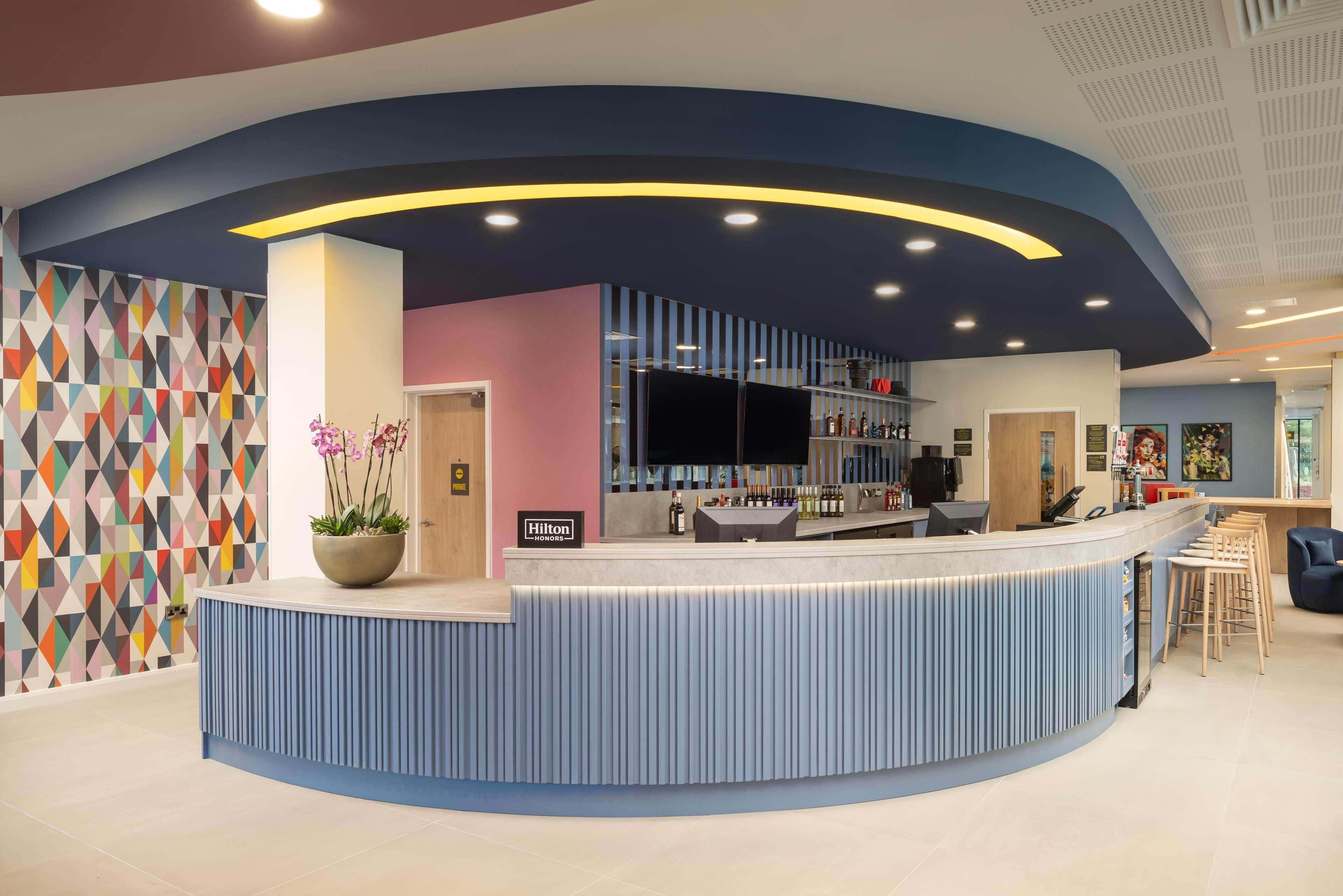 A modern hotel reception area with a curved blue counter, a potted plant, and a colorful geometric wall.