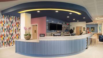 A modern hotel reception area with a curved blue counter, a potted plant, and a colorful geometric wall.