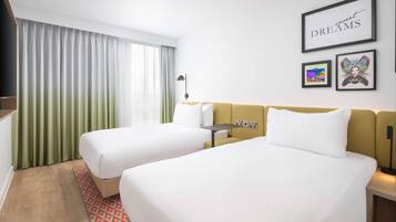 A hotel room with two beds, a desk, and a large window with curtains. There are framed pictures on the wall and a sign that says "sweet DREAMS".