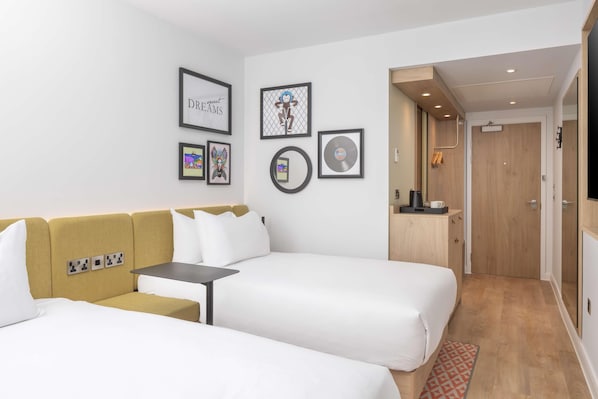 A hotel room with two beds, a sofa, a small table, and a wooden cabinet. There are framed pictures on the wall and a door leading to a bathroom. - Hampton By Hilton Nuneaton (Nuneaton)