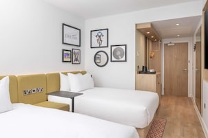 Habitación de hotel con dos camas, un sofá, una mesita y un armario de madera. En la pared hay cuadros enmarcados y una puerta que da a un baño.
