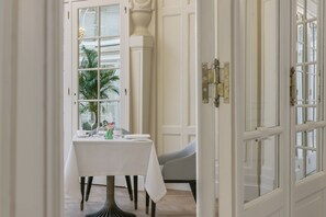 Un coin repas avec une table blanche dressée pour deux personnes, une chaise et une vue sur un palmier à travers une porte vitrée.