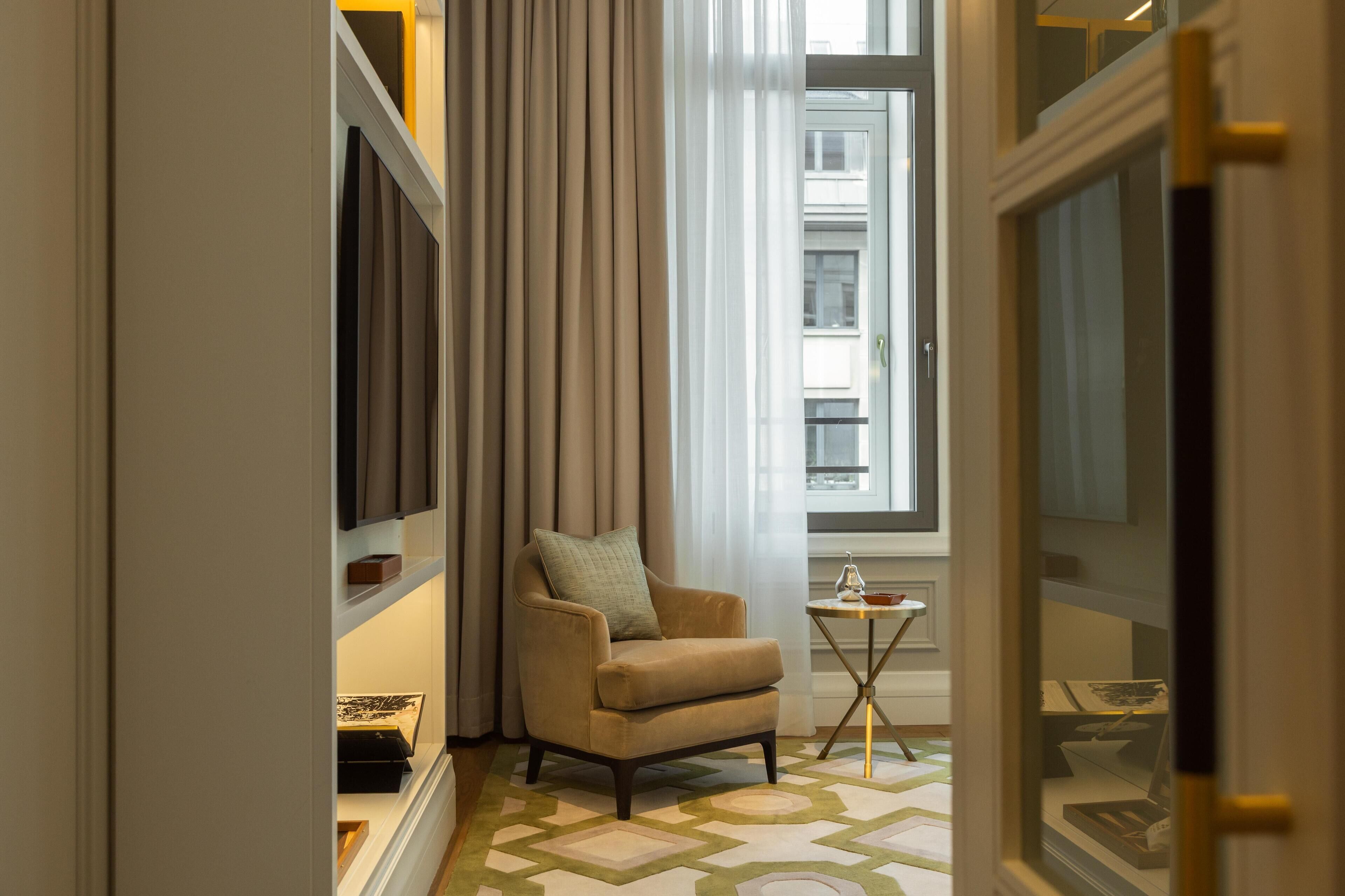 Une chambre d’hôtel avec un fauteuil beige, une petite table avec un verre et une fenêtre avec des rideaux.