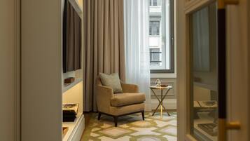 Une chambre d’hôtel avec un fauteuil beige, une petite table avec un verre et une fenêtre avec des rideaux.