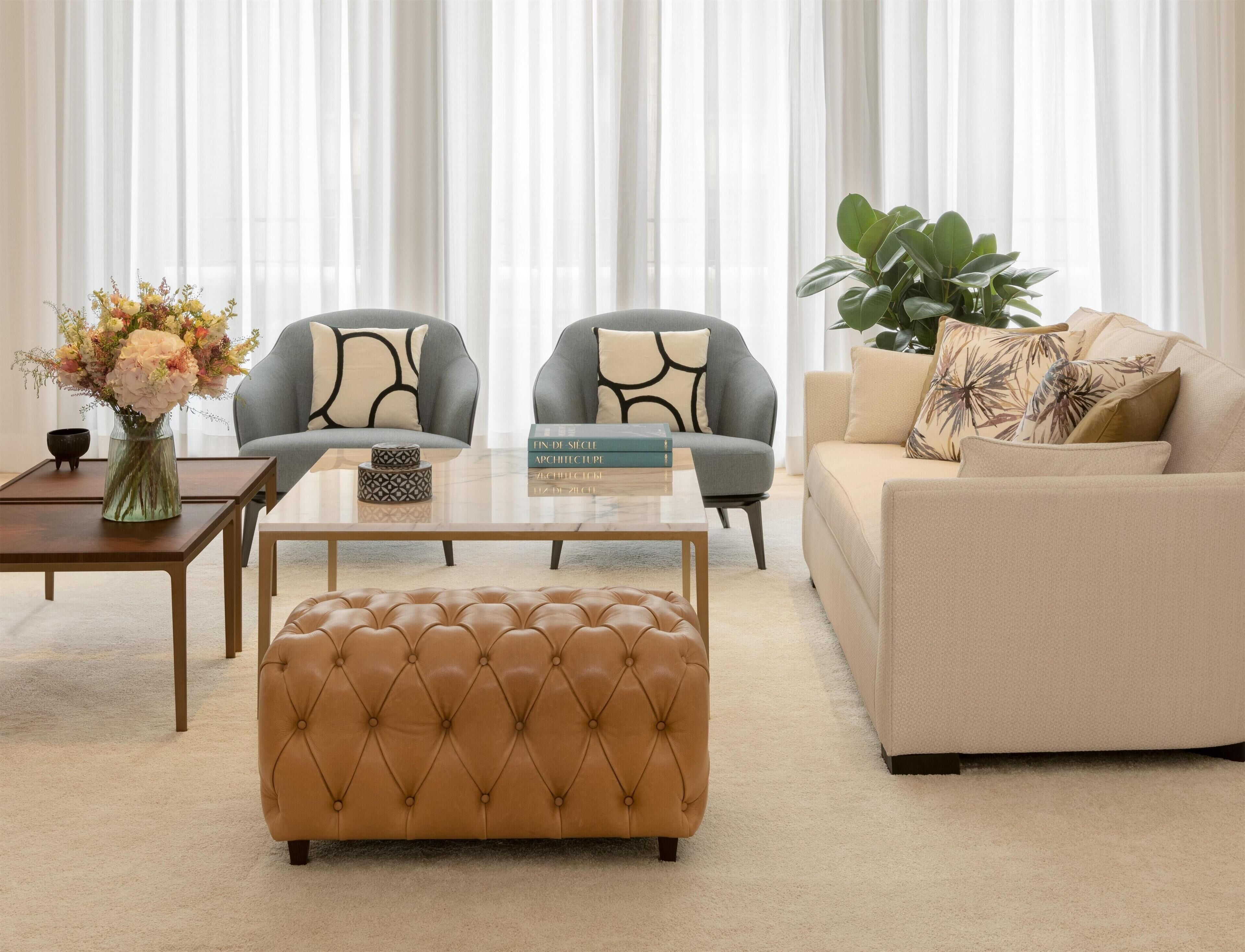 Un salon avec un canapé beige, deux fauteuils gris, une table basse en verre, un pouf marron, un vase de fleurs et une bibliothèque remplie de livres.