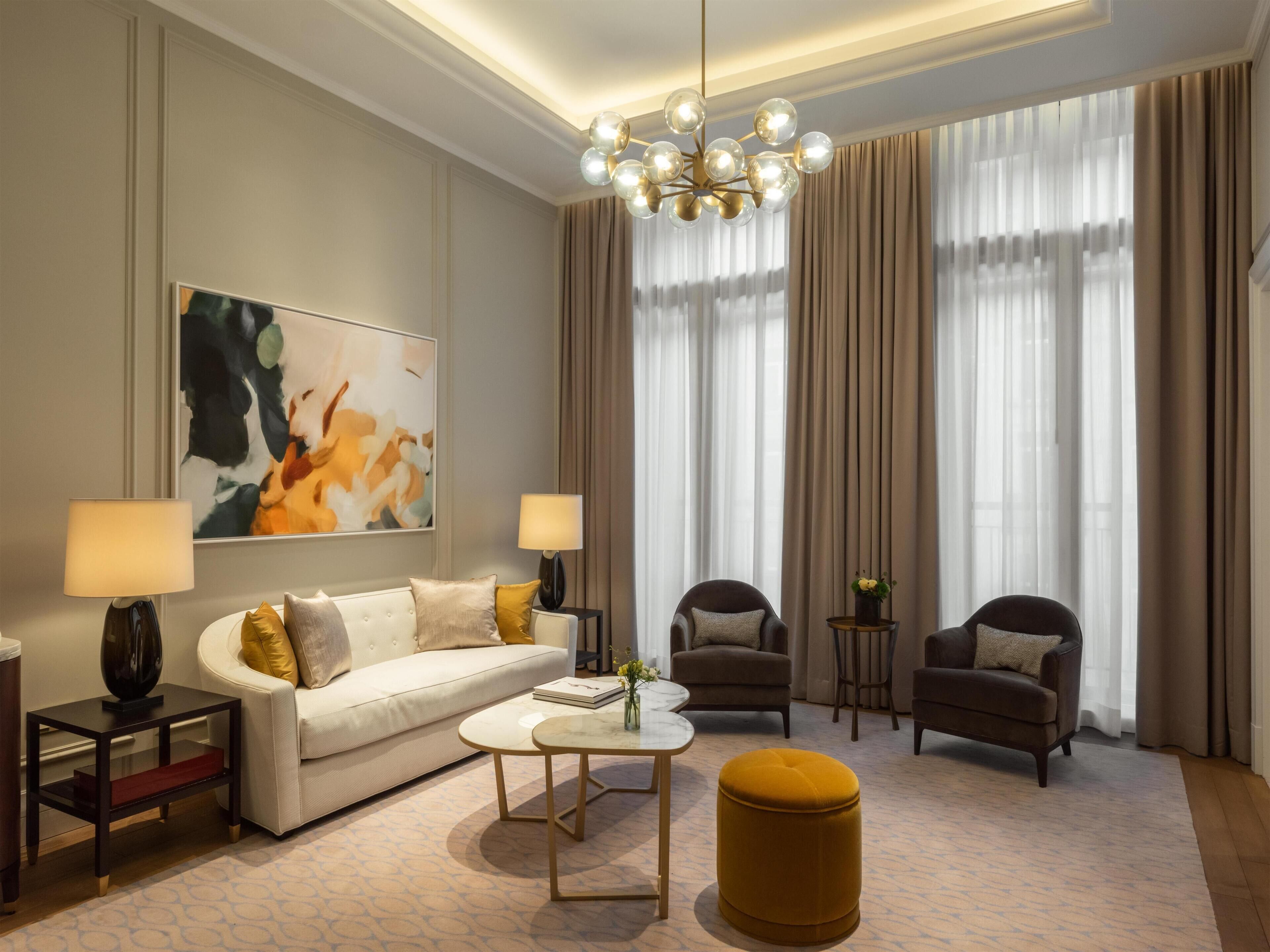Una sala de estar moderna con un sofá, dos sillones, una mesa de centro y un otoman redondos. Un candelabro cuelga del techo, y hay un cuadro abstracto grande en la pared.