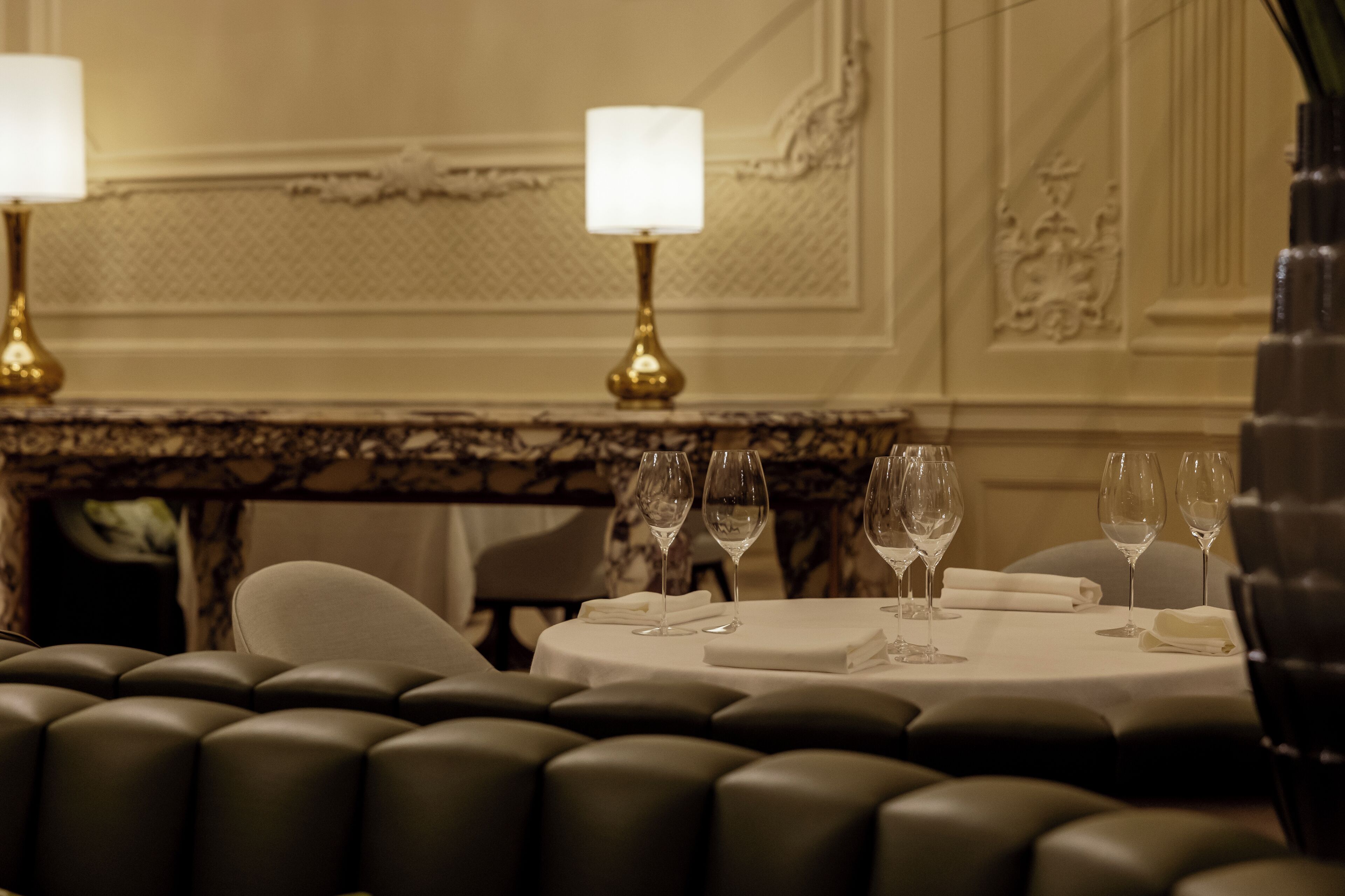 A dining table with four wine glasses and a folded napkin, set against a marble wall with decorative molding.